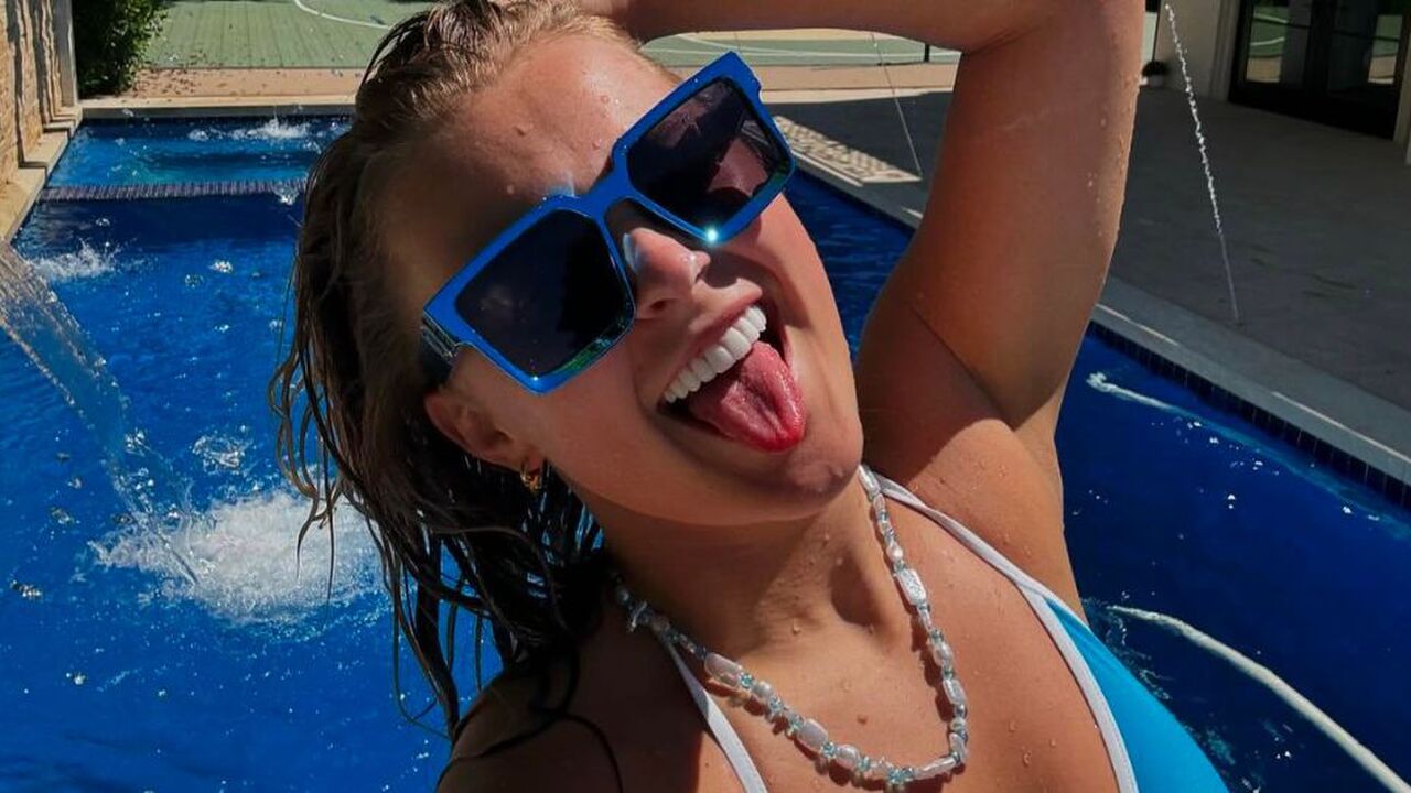 Selfie van JoJo Siwa waarin ze haar tong uitsteekt, een grote blauwe zonnebril op heeft en voor een zwembad staat.